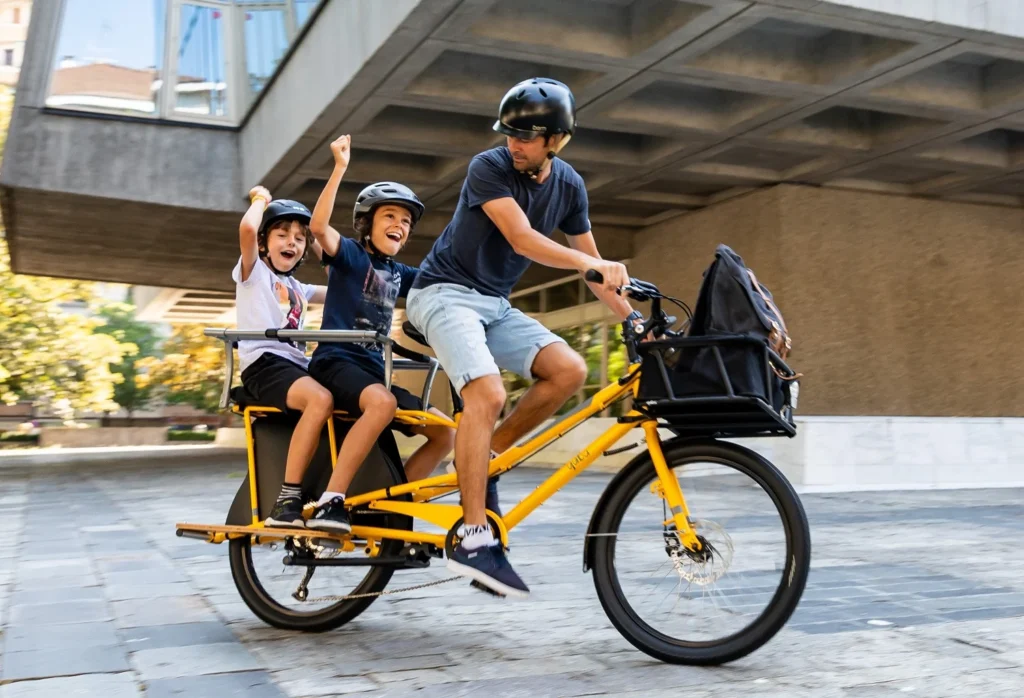 Padre disfrutando con sus dos hijos pequeños mientas montan en una bicicleta de carga de tipo longtail en la ciudad